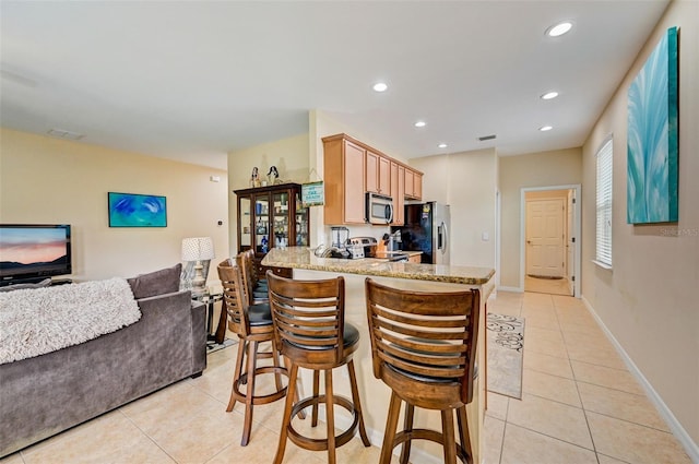 kitchen with light brown cabinets, kitchen peninsula, stainless steel appliances, light tile flooring, and a kitchen breakfast bar