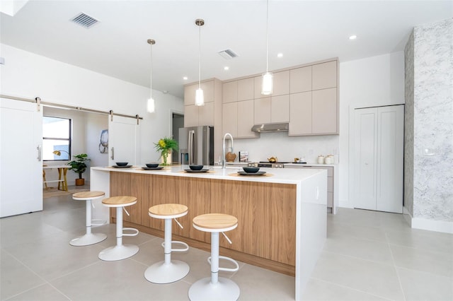 kitchen featuring a barn door, light countertops, an island with sink, stainless steel fridge, and decorative light fixtures