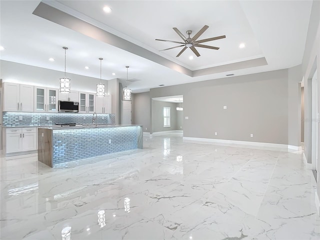 kitchen with a tray ceiling, a kitchen island with sink, ceiling fan, decorative light fixtures, and white cabinetry