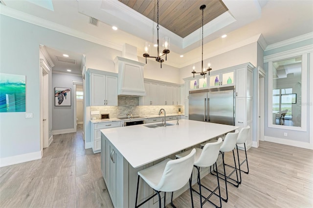 kitchen featuring a kitchen island with sink, built in fridge, sink, a tray ceiling, and decorative light fixtures