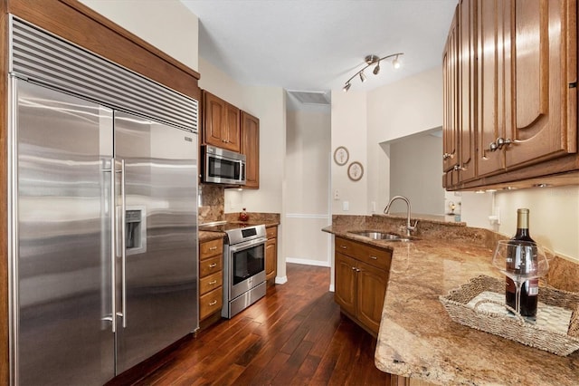 kitchen featuring light stone counters, dark wood finished floors, appliances with stainless steel finishes, brown cabinetry, and a sink