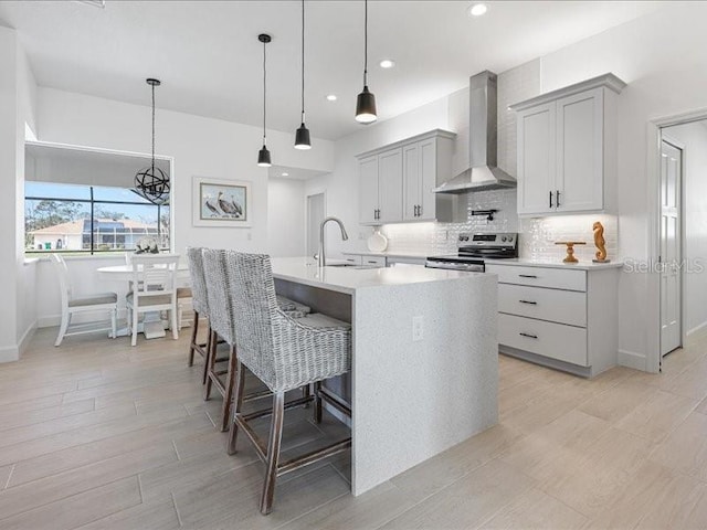 kitchen with electric stove, pendant lighting, a kitchen island with sink, backsplash, and wall chimney exhaust hood