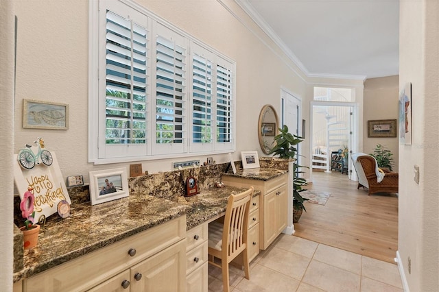 kitchen with light brown cabinets, ornamental molding, dark stone counters, built in desk, and light tile patterned flooring