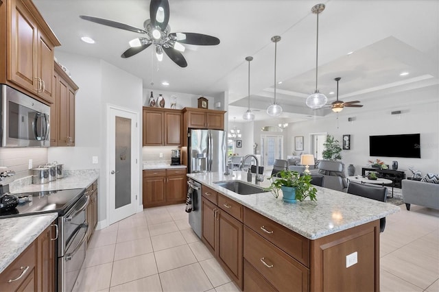 kitchen with a center island with sink, appliances with stainless steel finishes, brown cabinets, a tray ceiling, and a sink