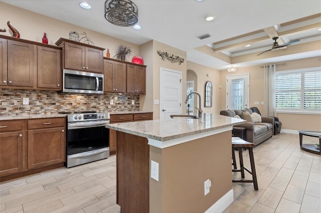 kitchen with coffered ceiling, appliances with stainless steel finishes, a kitchen island with sink, backsplash, and ceiling fan