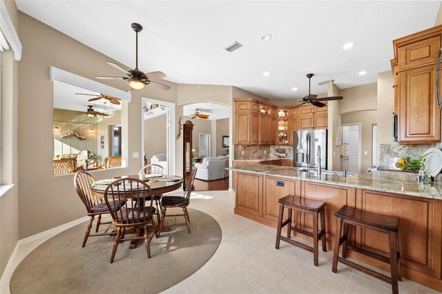 kitchen featuring a peninsula, stainless steel refrigerator with ice dispenser, brown cabinets, light stone countertops, and tasteful backsplash