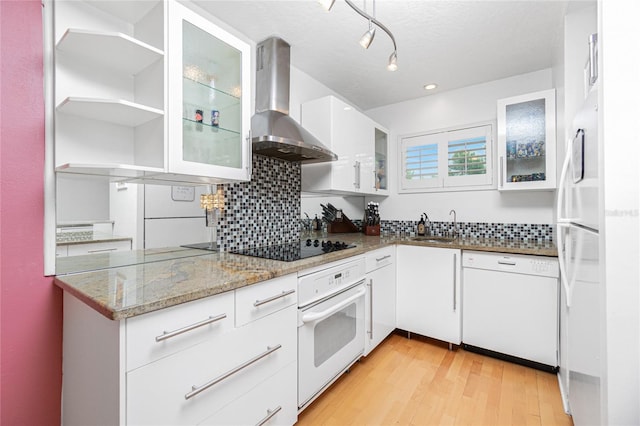 kitchen with white appliances, wall chimney range hood, sink, light stone counters, and white cabinetry