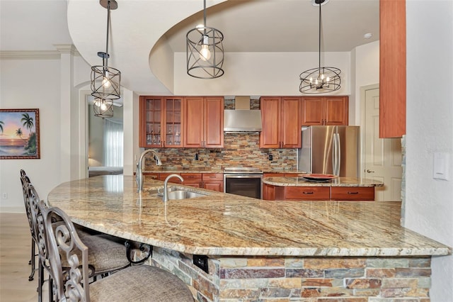 kitchen featuring brown cabinets, a sink, appliances with stainless steel finishes, wall chimney range hood, and decorative backsplash