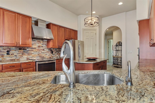 kitchen with appliances with stainless steel finishes, light stone countertops, a chandelier, wall chimney exhaust hood, and decorative backsplash