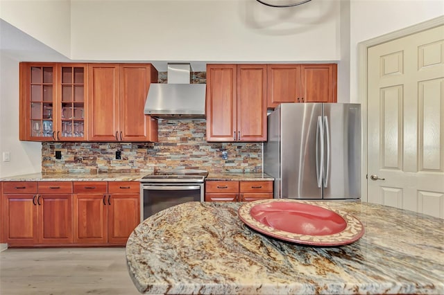 kitchen featuring light stone counters, glass insert cabinets, appliances with stainless steel finishes, wall chimney range hood, and decorative backsplash