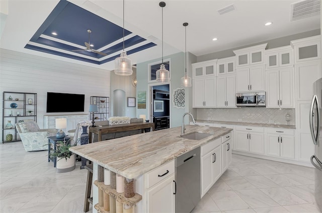 kitchen featuring appliances with stainless steel finishes, white cabinets, a tray ceiling, a kitchen island with sink, and sink