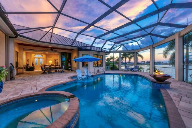 pool at dusk featuring a patio, glass enclosure, a water view, an in ground hot tub, and ceiling fan