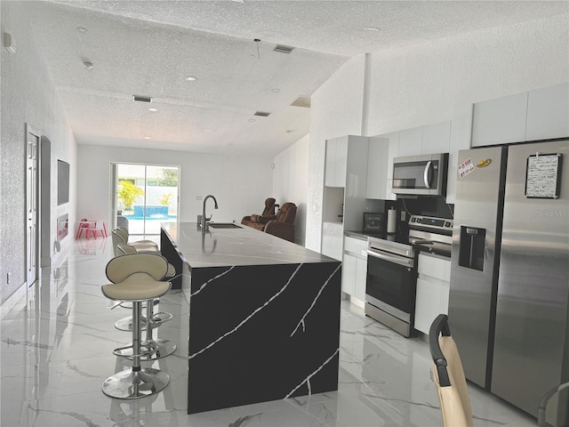 kitchen featuring appliances with stainless steel finishes, white cabinetry, a textured ceiling, a center island with sink, and sink