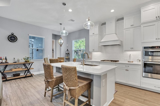 kitchen featuring hanging light fixtures, a center island with sink, white cabinets, premium range hood, and sink