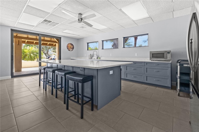 kitchen with a drop ceiling, a center island with sink, a kitchen breakfast bar, ceiling fan, and appliances with stainless steel finishes