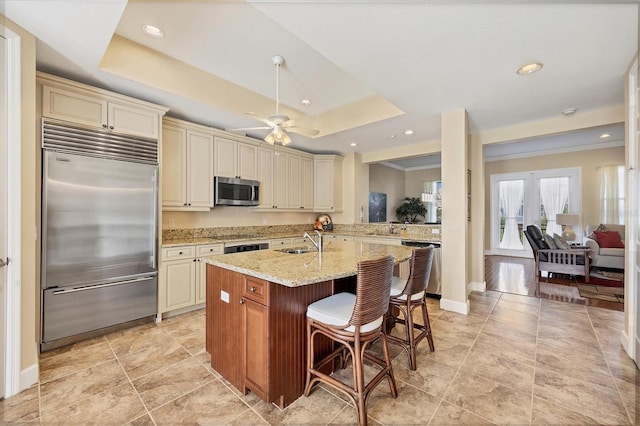 kitchen with appliances with stainless steel finishes, a kitchen breakfast bar, a tray ceiling, light stone countertops, and cream cabinetry