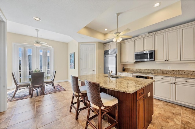 kitchen with a breakfast bar, a kitchen island with sink, light stone counters, a tray ceiling, and stainless steel appliances