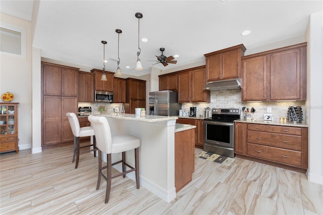 kitchen featuring an island with sink, hanging light fixtures, stainless steel appliances, backsplash, and a kitchen bar