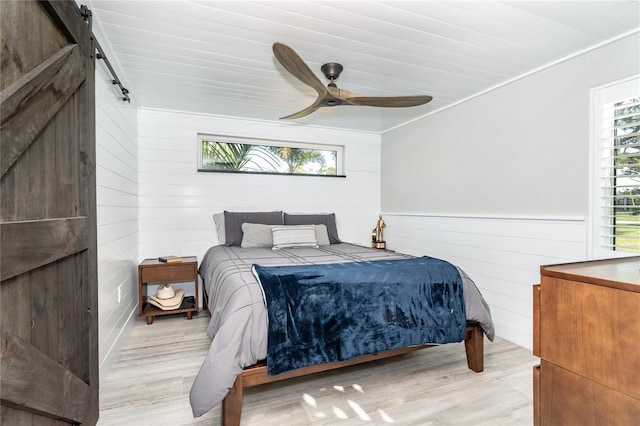 bedroom featuring ceiling fan, a barn door, light wood-type flooring, crown molding, and wooden walls