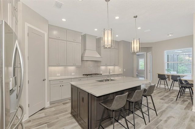 kitchen with stainless steel appliances, a center island with sink, sink, custom exhaust hood, and decorative light fixtures