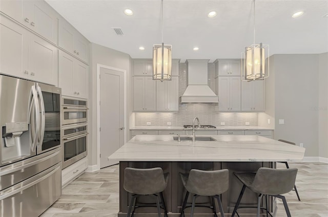 kitchen featuring stainless steel appliances, custom range hood, pendant lighting, light stone countertops, and a kitchen island with sink