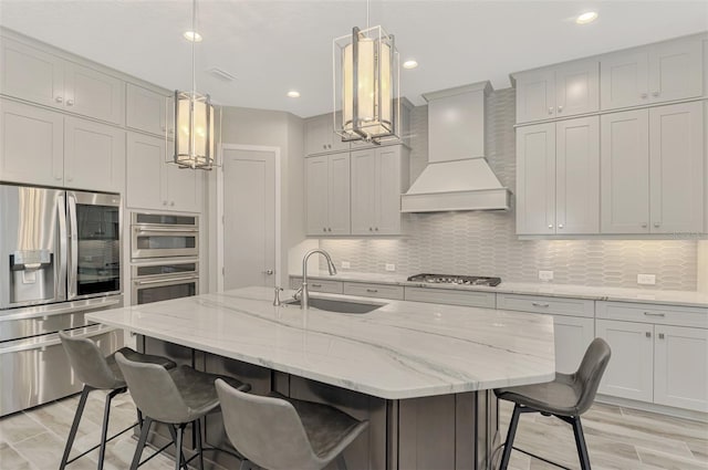 kitchen with sink, a kitchen island with sink, custom range hood, appliances with stainless steel finishes, and decorative light fixtures