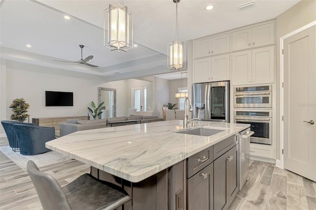 kitchen featuring stainless steel appliances, light stone countertops, ceiling fan, hanging light fixtures, and an island with sink