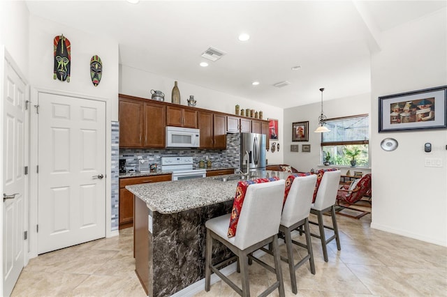 kitchen featuring a kitchen bar, an island with sink, light stone countertops, pendant lighting, and white appliances