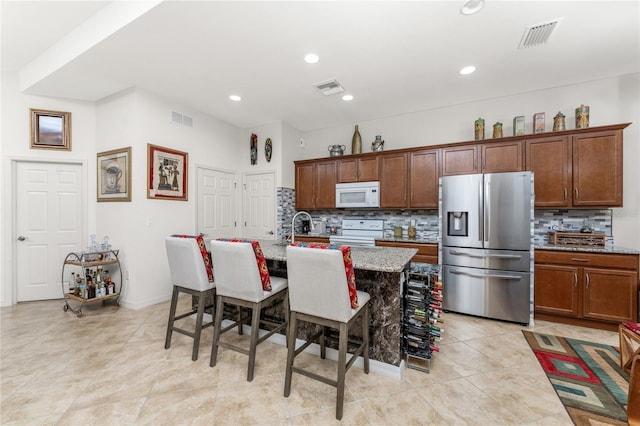 kitchen featuring light stone counters, decorative backsplash, a breakfast bar area, a kitchen island with sink, and white appliances