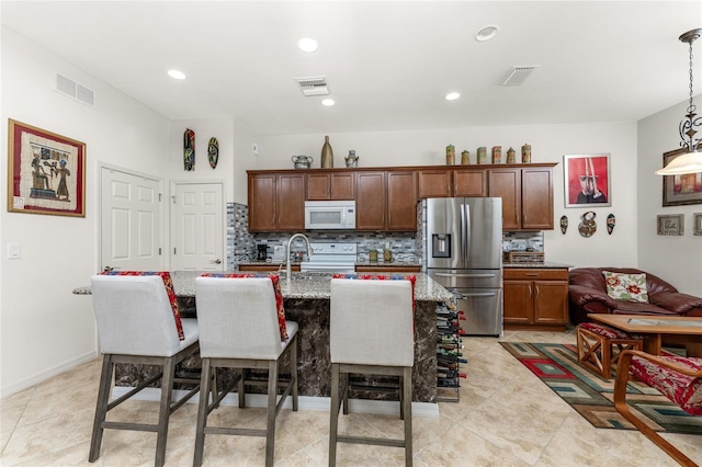 kitchen featuring light stone counters, a kitchen breakfast bar, white appliances, a kitchen island with sink, and pendant lighting