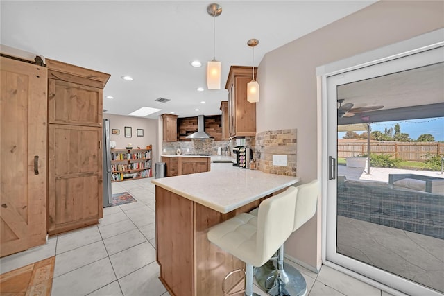 kitchen featuring a barn door, kitchen peninsula, hanging light fixtures, wall chimney exhaust hood, and decorative backsplash