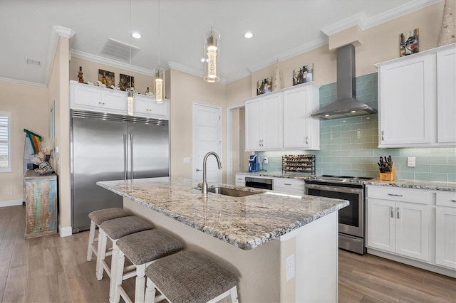 kitchen with wall chimney exhaust hood, an island with sink, white cabinetry, light wood-type flooring, and appliances with stainless steel finishes