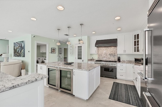 kitchen featuring high quality appliances, a center island with sink, hanging light fixtures, wall chimney exhaust hood, and beverage cooler