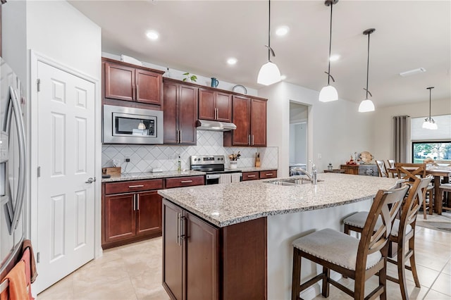 kitchen with light stone countertops, appliances with stainless steel finishes, a kitchen island with sink, decorative light fixtures, and a breakfast bar area