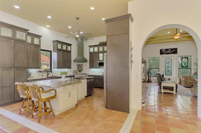 kitchen with a center island with sink, wall chimney range hood, decorative backsplash, light stone countertops, and light tile patterned flooring