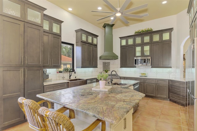 kitchen with appliances with stainless steel finishes, light stone counters, wall chimney range hood, sink, and an island with sink