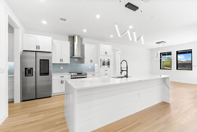 kitchen with wall chimney exhaust hood, hanging light fixtures, a kitchen island with sink, white cabinets, and appliances with stainless steel finishes