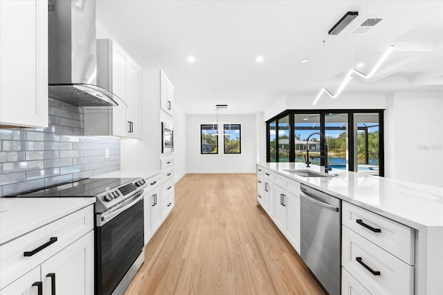 kitchen with wall chimney exhaust hood, stainless steel appliances, decorative light fixtures, white cabinets, and light wood-type flooring