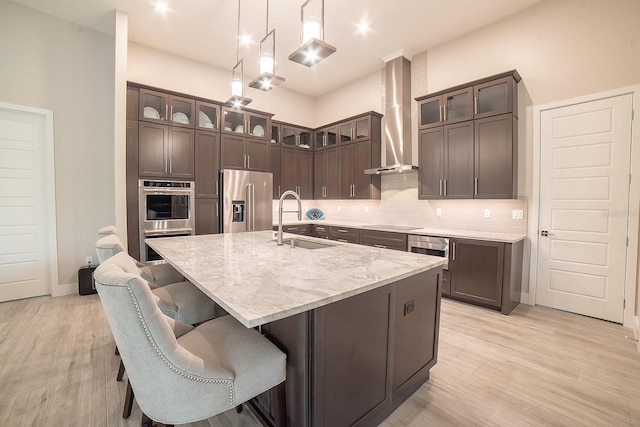 kitchen featuring appliances with stainless steel finishes, wall chimney exhaust hood, a kitchen island with sink, sink, and hanging light fixtures