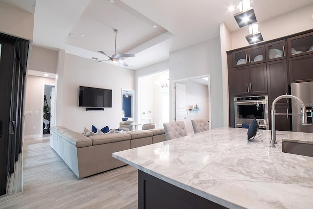 kitchen with oven, a raised ceiling, ceiling fan, light stone counters, and dark brown cabinetry