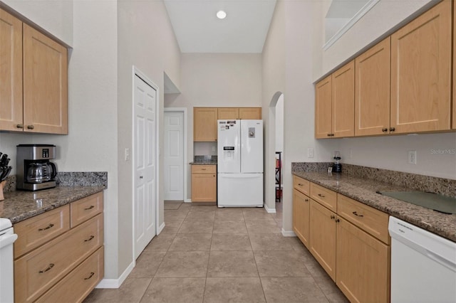 kitchen featuring a towering ceiling, white appliances, light brown cabinets, light tile patterned floors, and dark stone countertops