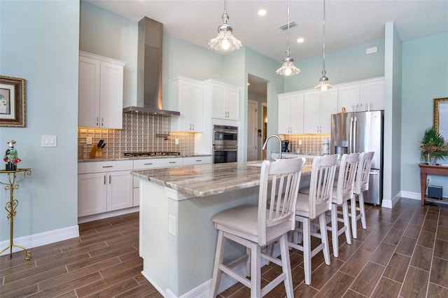 kitchen featuring white cabinetry, wall chimney exhaust hood, an island with sink, pendant lighting, and appliances with stainless steel finishes