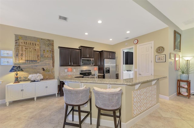 kitchen with a kitchen bar, decorative backsplash, light stone countertops, dark brown cabinetry, and stainless steel appliances