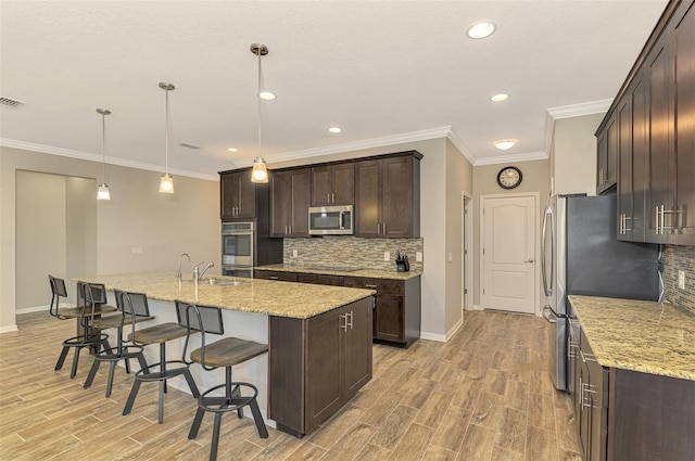 kitchen featuring appliances with stainless steel finishes, dark brown cabinets, pendant lighting, a center island with sink, and light hardwood / wood-style flooring