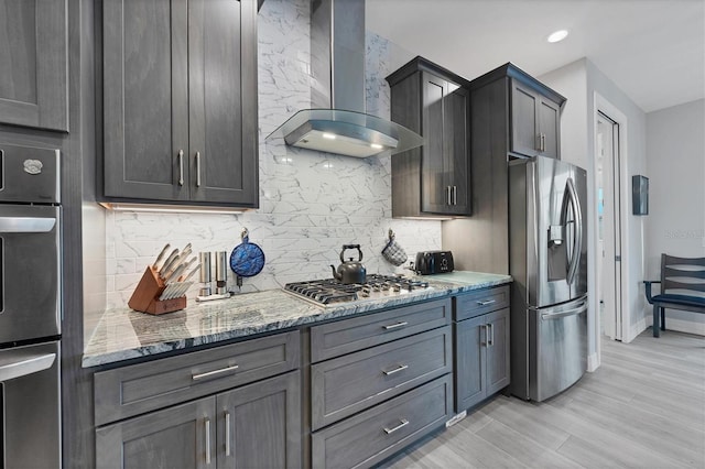 kitchen with tasteful backsplash, wall chimney exhaust hood, stainless steel appliances, dark stone countertops, and light hardwood / wood-style floors