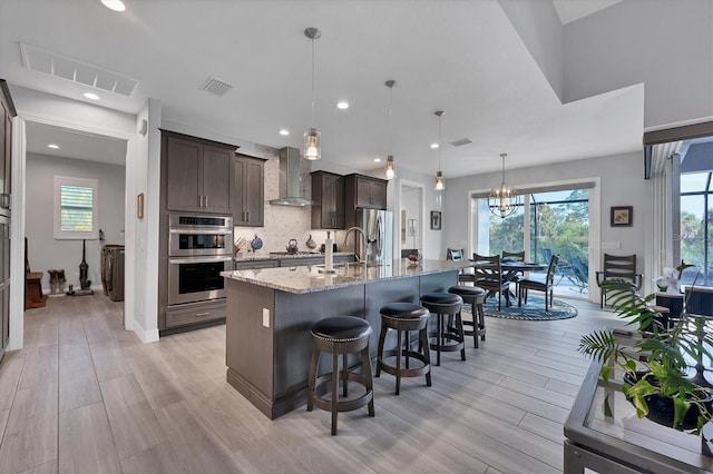 kitchen with light hardwood / wood-style floors, wall chimney range hood, a kitchen island with sink, and a wealth of natural light