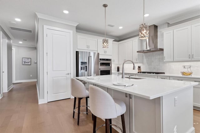 kitchen featuring stainless steel appliances, white cabinetry, a kitchen island with sink, and wall chimney range hood