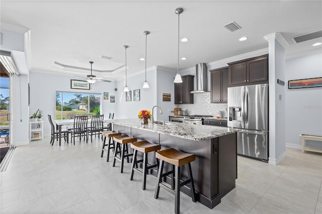 kitchen with a kitchen island with sink, wall chimney exhaust hood, ceiling fan, appliances with stainless steel finishes, and dark brown cabinets