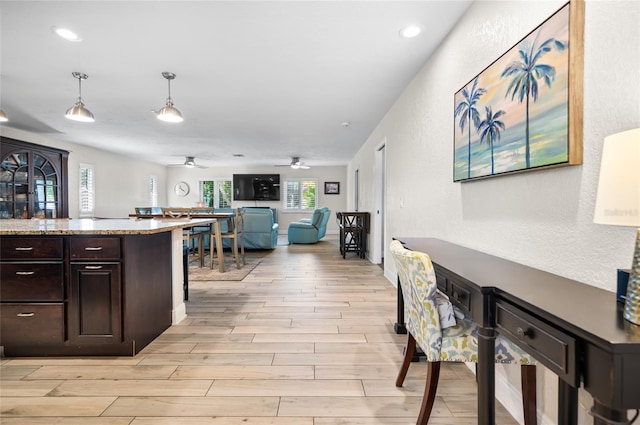 kitchen with light stone counters, dark brown cabinetry, ceiling fan, pendant lighting, and light hardwood / wood-style floors