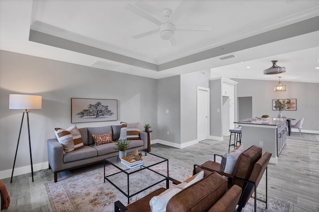 living room featuring a tray ceiling, crown molding, ceiling fan with notable chandelier, and light wood-type flooring